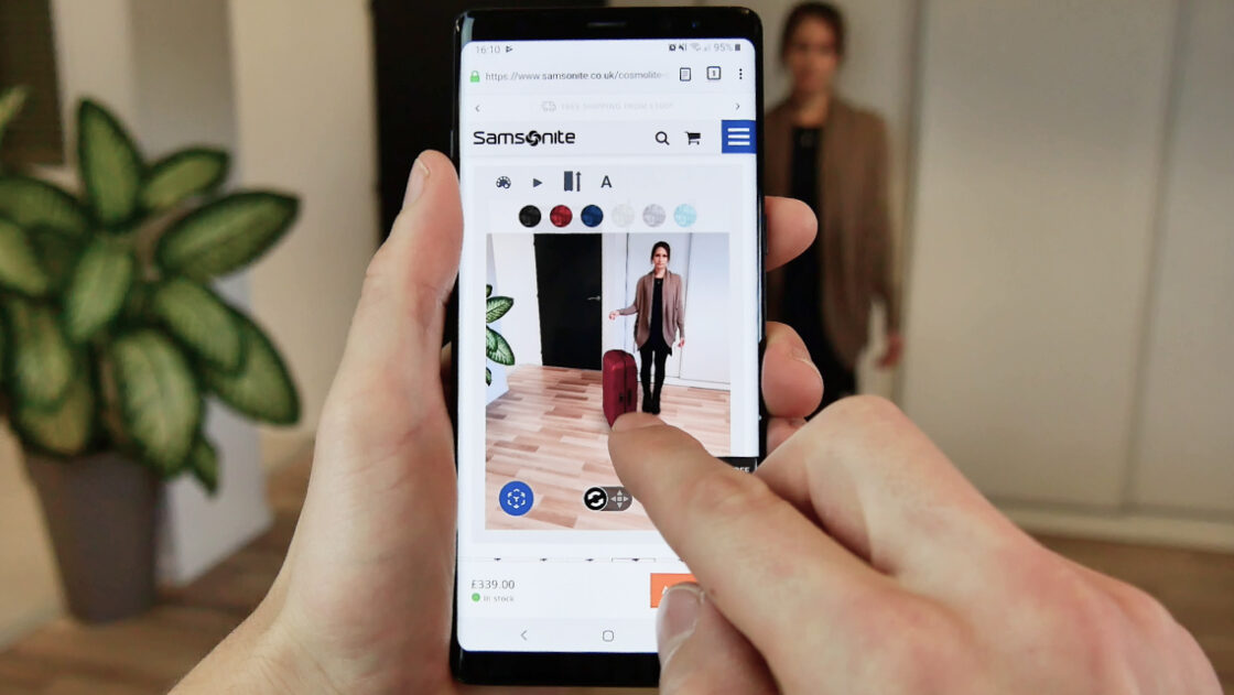 Screenshot of the deep red Samsonite Lite-box being tryed-out in augmented reality by female customer in her own interior. Second customer is holding the smartphone and is able to switch between all available colors and sizes directly from the product page.