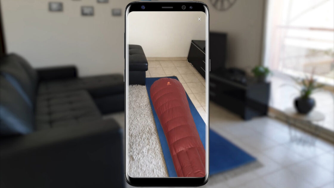 Customer trying the Deuter Astro pro 800 sleeping bag in augmented reality on their living room floor, using a mobile device. Sleeping bag is a burgundy color.