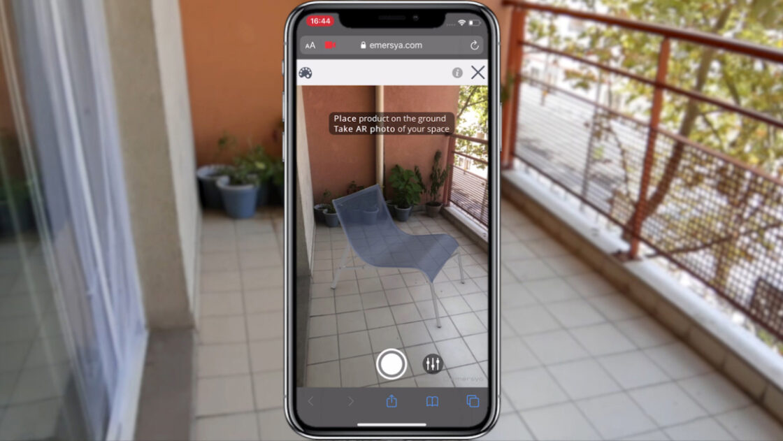 Customer trying out an Alias arm chair to scale on their balcony thanks to the smartphone's camera. Smartphone screen in the middle of the image with real-life scenery in the background.