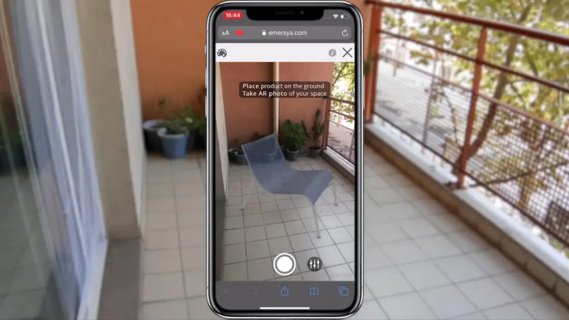 Customer trying out an Alias arm chair to scale on their balcony thanks to the smartphone's camera. Smartphone screen in the middle of the image with real-life scenery in the background.