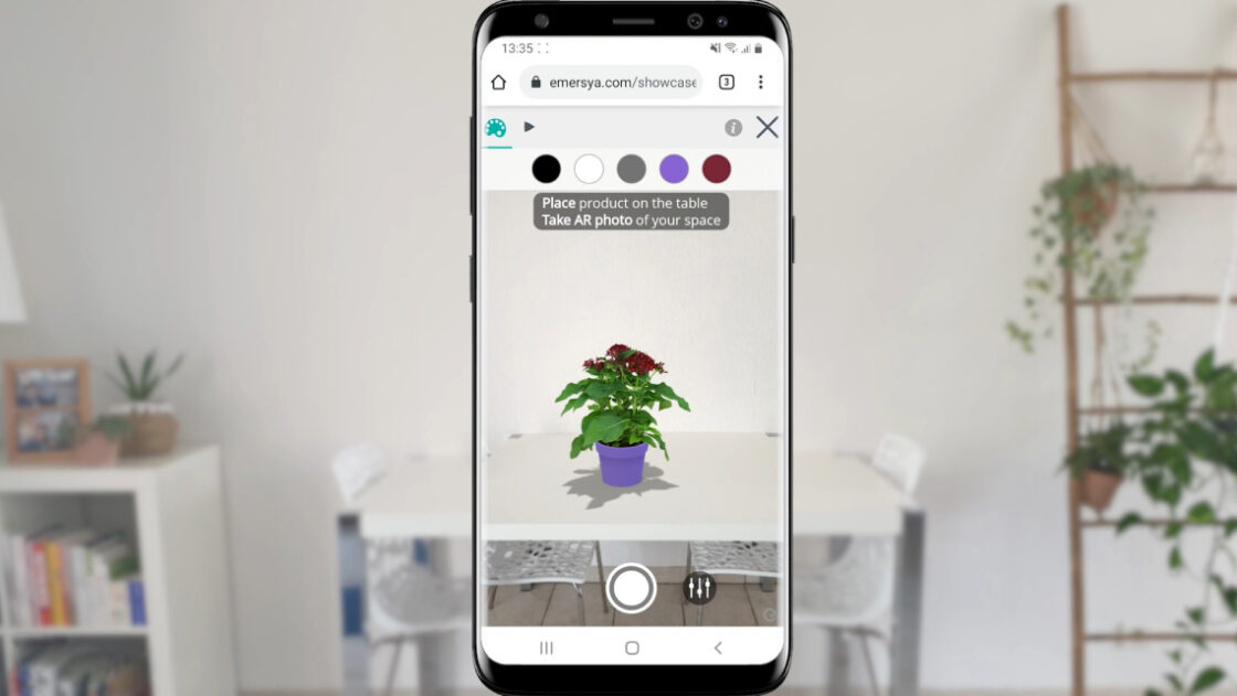 Smartphone screen in the middle of the image, real-life scenery in the background. Plant pot displayed in AR on a tabletop : red pentas, violet plant pot. Customers can preview all the available colorways for the flower pot directly in the AR view (black, white, grey, violet, red)