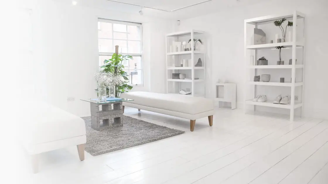 Modern living room on an apartment with wooden parquet flooring and a large sash window along the far wall. There are 2 large shelving units with decorations on them against the back wall and a grey shag rug i the middle with a square glass coffee table on it. Either side of the rug are two long white upholstered ottoman bench seats.